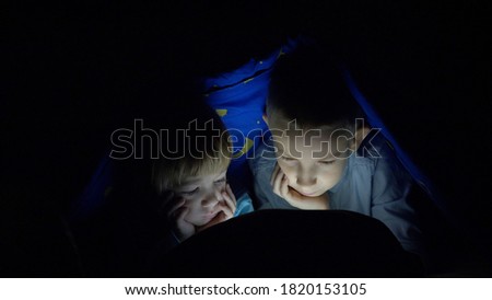 Children hold head in hands and look at tablet screen under blanket, bedtime 