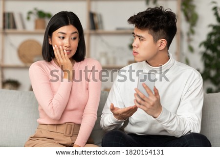 Infidelity. Jealous Korean Husband Catching Wife On Cheating, Showing Her Phone Sitting On Sofa At Home. Boyfriend Suspecting Affair. Jealousy And Relationship Problems Concept Royalty-Free Stock Photo #1820154131