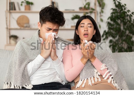 Sick Asian Family Couple Sneezing Blowing Nose In Paper Napkin Having Flu Cold Sitting On Sofa At Home. Seasonal Allergy And Rhinitis Illness. Ill Chinese Spouses On Self-Isolation Concept. Royalty-Free Stock Photo #1820154134