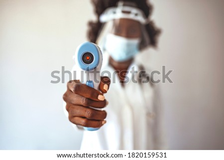 Woman doctor in safety face mask and white coat holding infrared thermometer, standing isolated on white background. Check body temperature for covid-19. Shot of a doctor using an infrared thermometer