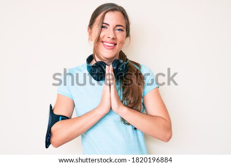 Beautiful young caucasian woman wearing gym clothes and using headphones praying with hands together asking for forgiveness smiling confident. 