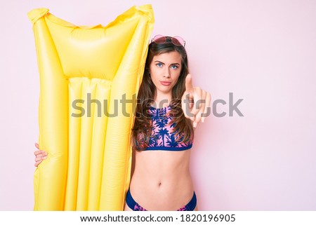 Beautiful young caucasian woman wearing bikini and holding summer matress float pointing with finger to the camera and to you, confident gesture looking serious 
