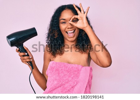 Young african american woman wearing shower towel holding dryer smiling happy doing ok sign with hand on eye looking through fingers 