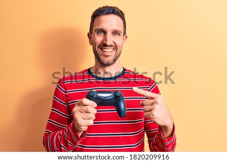 Young handsome gamer man playing video game using gamepad over yellow background smiling happy pointing with hand and finger