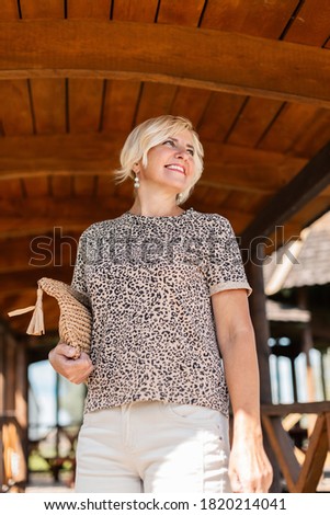 Beautiful fashionable elderly woman with a smile in a leopard print t-shirt with a woven handbag resting on vacation on summer day off