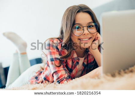 Mirthful teenager wearing glasses lying on the bed in front of a laptop with her hand touching the cheek Royalty-Free Stock Photo #1820216354