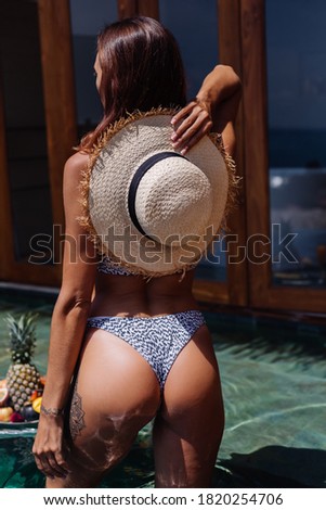 Beautiful caucasian tanned tattooed woman in bikini and straw hat with floating breakfast at amazing luxury bali style villa at sunny day by swimming pool, tropical background.  