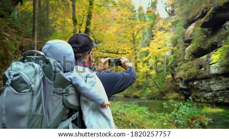 Male tourist with little son in touristic backpack  taking pictures of the autumn scenery by smartphone , Czech Switzerland, Bohemian Switzerland National Park Czechia 