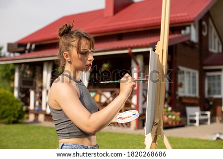 Young beautiful woman female artist painting picture on easel outside on loan near country house, outdoors relaxing activity, creative hobby, creation, inspiration, summer weekend recreation concept