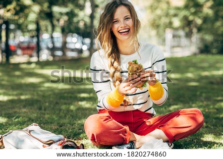  Happy student female sitting on the green grass at the college campus on a sunny day, have lunch and studying outdoors. A smiling young woman takes a rest eating fast food and learning in the park. Royalty-Free Stock Photo #1820274860