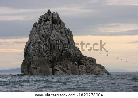 Epic seascape. Small figures of people are visible on a huge rock in the sea. These are sea hunters looking for sea animals (walrus and seals) to give a signal to hunt. Bering Strait, Chukotka, Russia