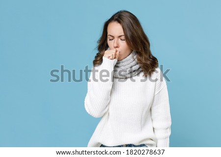 Sick young woman in white sweater gray scarf coughing covering mouth with hand keeping eyes closed isolated on blue background studio. Healthy lifestyle ill sick disease treatment cold season concept Royalty-Free Stock Photo #1820278637
