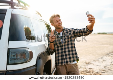 Attractive young man leaning on a car while standing at the beach, using mobile phone, taking a selfie