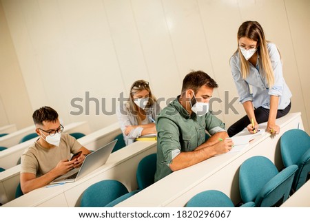 Young female instructor wearing face protective medical mask for virus protection and helping male student at lecture hall