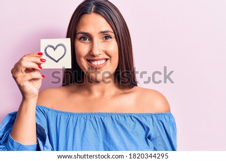 Young beautiful brunette woman holding reminder with heart looking positive and happy standing and smiling with a confident smile showing teeth 
