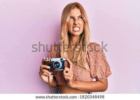 Beautiful blonde young woman holding vintage camera angry and mad screaming frustrated and furious, shouting with anger looking up. 