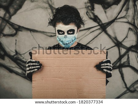 Happy Halloween. kid with medical mask in a skeleton costume holding brown board over gray background