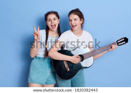 Happy women friends with guitar and showing v sign, keeping mouths opened, wearing white t shirt and jeans shorts, posing isolated over blue background, happy females, singing songs.