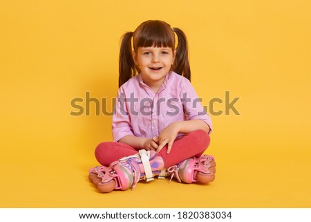 Little charming cute girl with roller skates sitting on floor with crossed legs, looks at camera, female child with two ponytail isolated over yellow background.