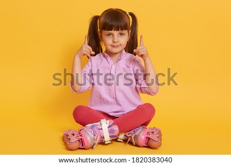 Dark haired little girl wearing roller skates, shirt and leggins, looking directly at camera and pointing up with fore fingers, sitting with crossed legs against yellow wall.