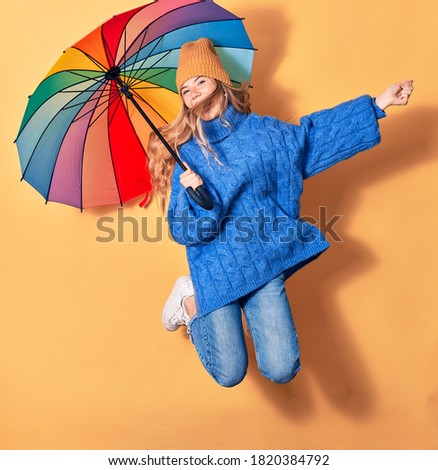 Young beautiful woman wearing winter clothes smiling happy. Jumping with smile on face holding colorful umbrella over isolated yellow background