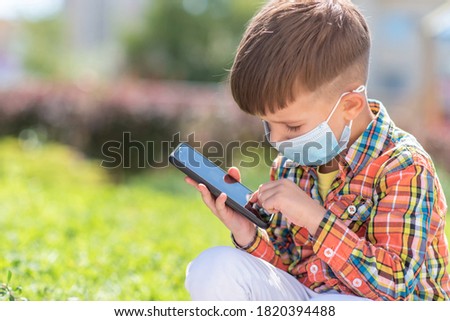 A kid in a medical mask sits on the grass and looks in the phone cartoons in the summer at sunset. Child with a mobile phone in his hands. Prevention against coronavirus Covid-19 during a pandemic