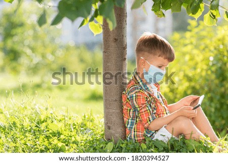 A child in a medical mask sits on the grass and looks at the phone in the summer at sunset. Boy with a mobile phone in his hand. Prevention against coronavirus Covid-19 during a pandemic