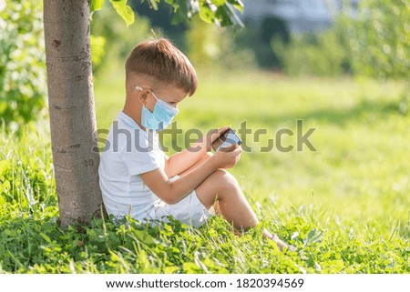 A child in a medical mask sits on the grass and looks at the phone in the summer at sunset. Boy with a mobile phone in his hand. Prevention against coronavirus Covid-19 during a pandemic