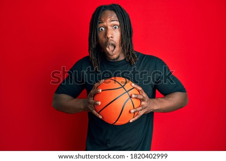 African american man with braids holding basketball ball afraid and shocked with surprise and amazed expression, fear and excited face. 