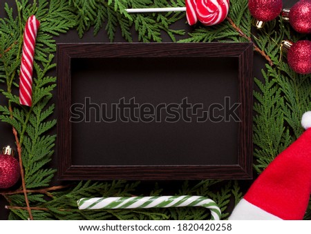 Christmas background with dark wooden picture frame, thuja branches, red baubles, lollipops and santa hat making the copy space