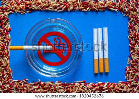 no smoking sign in a glass ashtray with a cigarette in it and cigarettes in a row next to, in a frame of broken matchsticks, on blue background