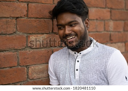 Closeup of a dark skinned young man posing beside old red brick wall. Relax profile picture with a confident smile on face | Indian dark skinned boy laughing