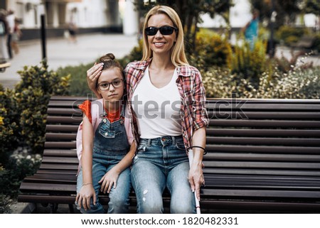Young blind mother sitting in schoolyard with her little daughter. They smiling and talking together.