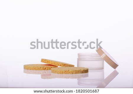 cosmetic cream jar and Three elastic Beige round sponge pads for face make-up cleaning reflected in glass surface isolated on white background. Daily, beauty care cosmetic. Face cream, eye cream