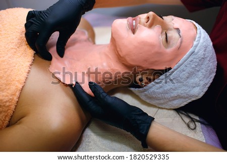 Brunette woman takes treatments in the spa. The pink mask was applied in a thick layer to the skin of the face. A master beautician takes care of the health of the client's skin
