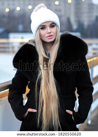 Blonde girl posing at the winter city in a black fur coat and a white hat