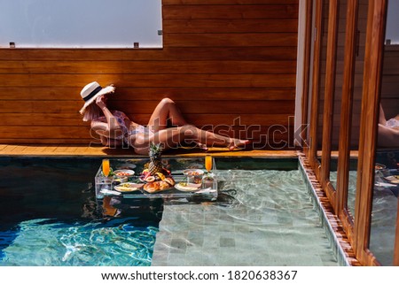 Fit pretty woman with perfect tanned bronze body skin lies on edge of pool  with floating breakfast on vacation in Thailand.