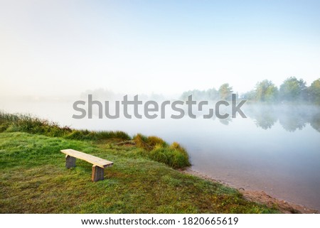 Green lake shore in a fog at sunrise. Old wooden bench close-up. Forest in the background. Idyllic rural scene. Atmospheric landscape. Early autumn in Europe. Pure nature, ecological resort, seasons