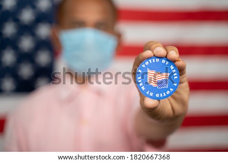 Man in medical mask showing I voted by mail sticker with US flag as background - Concept of mail in voting at USA election Royalty-Free Stock Photo #1820667368