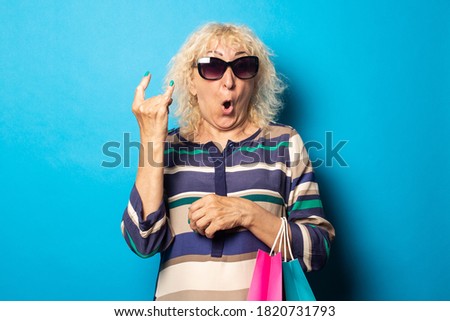 Old woman with glasses holds shopping bags and shows Rock and Roll gesture on blue background