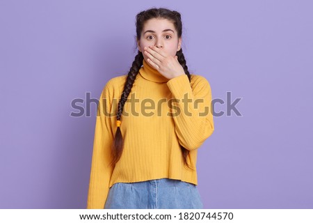 Young beautiful brunette woman wearing casual yellow sweater, posing over lilac background, being shocked, female covering mouth with hands for mistake.