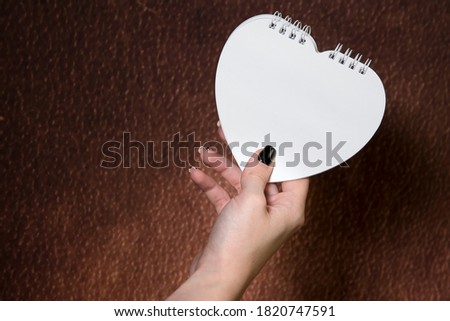 Close up of womanâ€™s hand holding white heart-shaped notepad, brown background, mock-up, empty space. Wedding stationery.