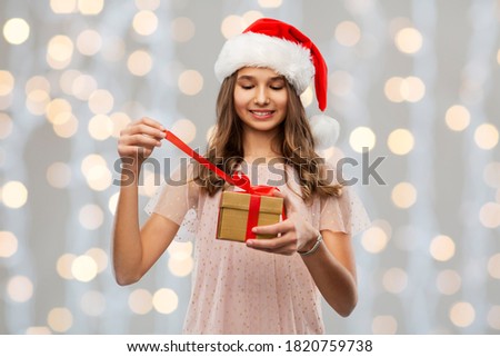 christmas, holidays and people concept - happy smiling teenage girl in santa helper hat opening gift box over festive lights background