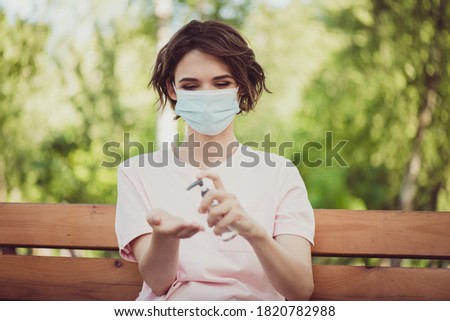 Photo of cute young lady relax sit bench wear cover face protective mask look down apply spray antiseptic alcohol liquid protect hands bacteria quarantine rules wear pink t-shirt park outside