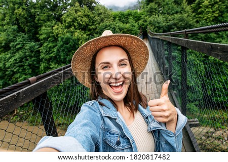 An attractive excited woman in straw hat stands on the bridge, holds a smartphone in hand and takes a selfie with a cheerful smile.