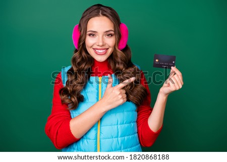 Happy client. Photo of cute charming young lady directing finger credit card offering fine banking service wear pink ear warmers blue vest red sweater isolated green color background