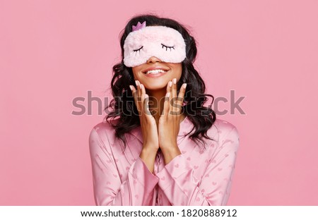 Delighted ethnic female in pajama and mask smiling and smearing lotion on face during skin care routine before sleep against pink background
 Royalty-Free Stock Photo #1820888912