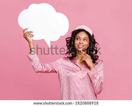Happy ethnic female in pajama smiling and looking at blank speech bubble against pink background
