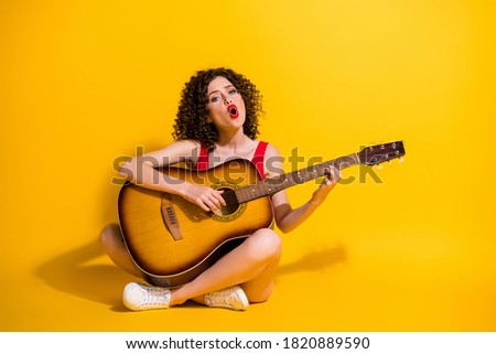 Portrait photo of hipster female musician with curly hair singing vocal voice song holding keeping playing melody guitar chords sitting down isolated on vivid yellow color background Royalty-Free Stock Photo #1820889590