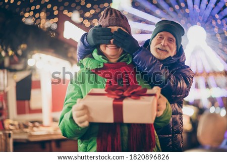 Photo of two people retired lovers couple palms cover eyes grandpa prepare giftbox grandma hold white box red bow wear coat red scarf headwear x-mas evening street park lights fair outside